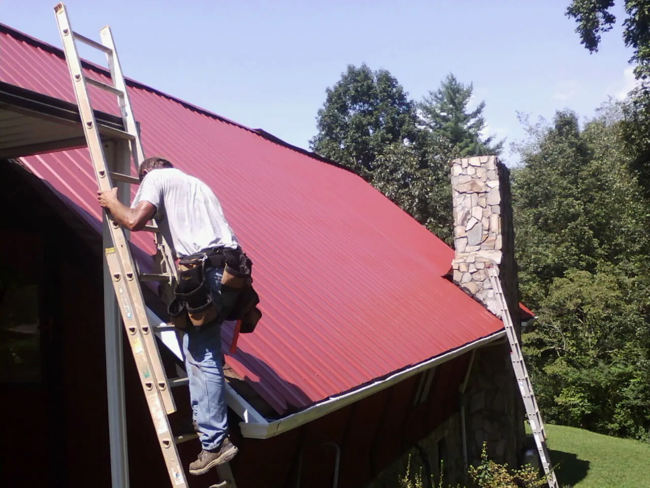 Roofer on a ladder beside a red metal roof and stone chimney — Soffit & Fascia Repair by Roofing Brothers in Swisshome